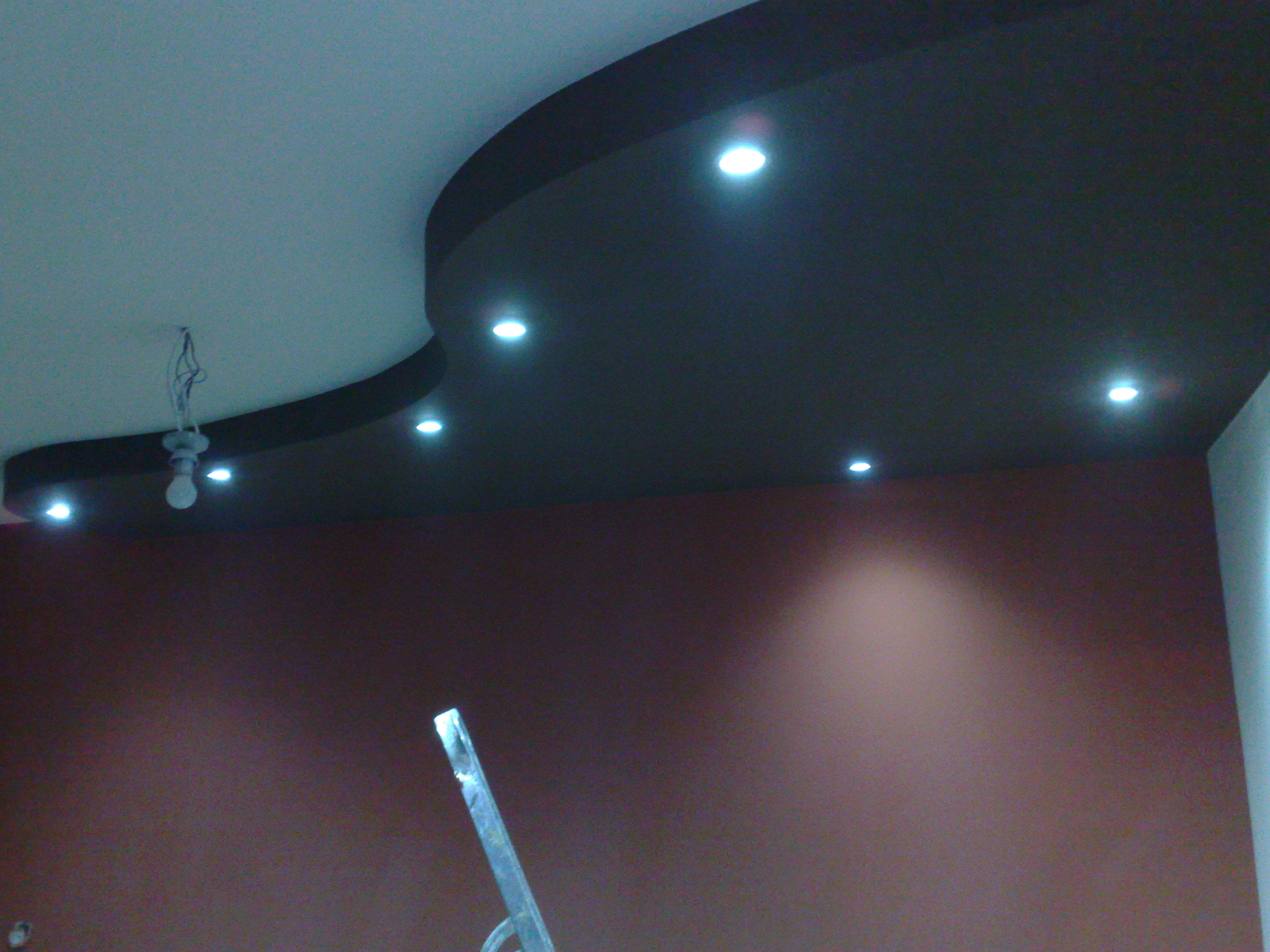 Wykończony sufit podwieszany z oświetleniem punktowym, widoczna drabina malarska i fragment żarówki na kablach.